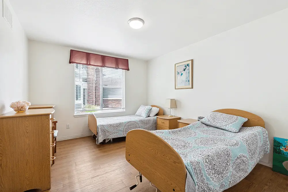 A clean, well-lit bedroom with two single beds, matching bedding, wooden furniture, a window with blinds and valance, and a framed picture on the wall.