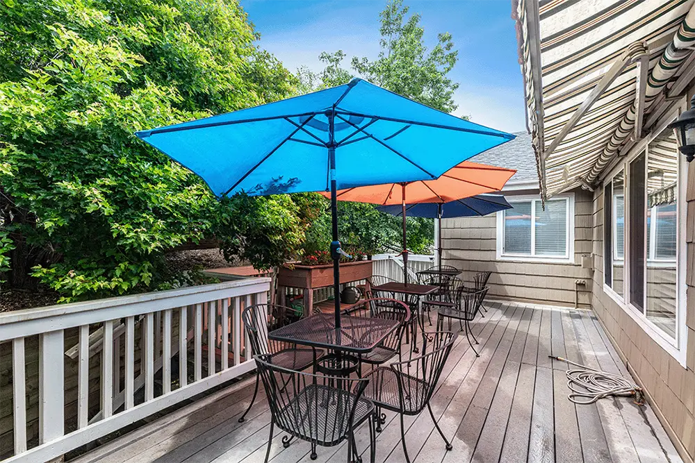 Outdoor wooden deck with black metal tables and chairs, shaded by blue and orange umbrellas, next to a beige house with windows and trees in the background.
