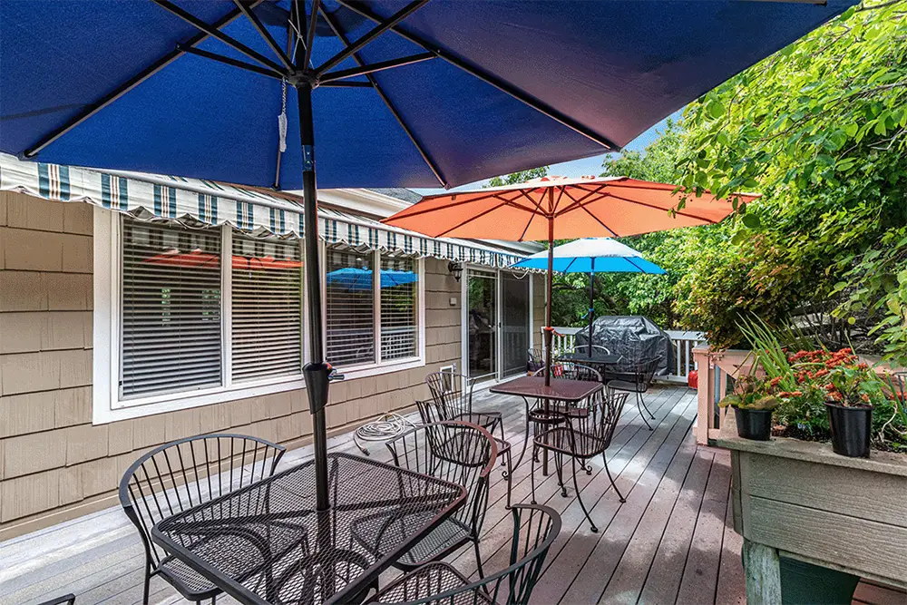 Outdoor patio with metal tables and chairs under blue and orange umbrellas, beside a house with sliding glass doors and striped awning, surrounded by greenery and potted plants.