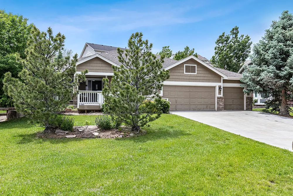 Single-story house with a three-car garage, front porch, and landscaped yard with trees and grass under a clear blue sky.