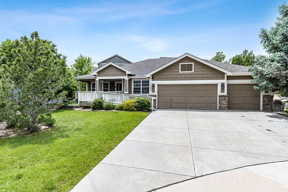 Single-story suburban house with a large driveway, three-car garage, front porch, and landscaped yard with trees and shrubs on a sunny day.