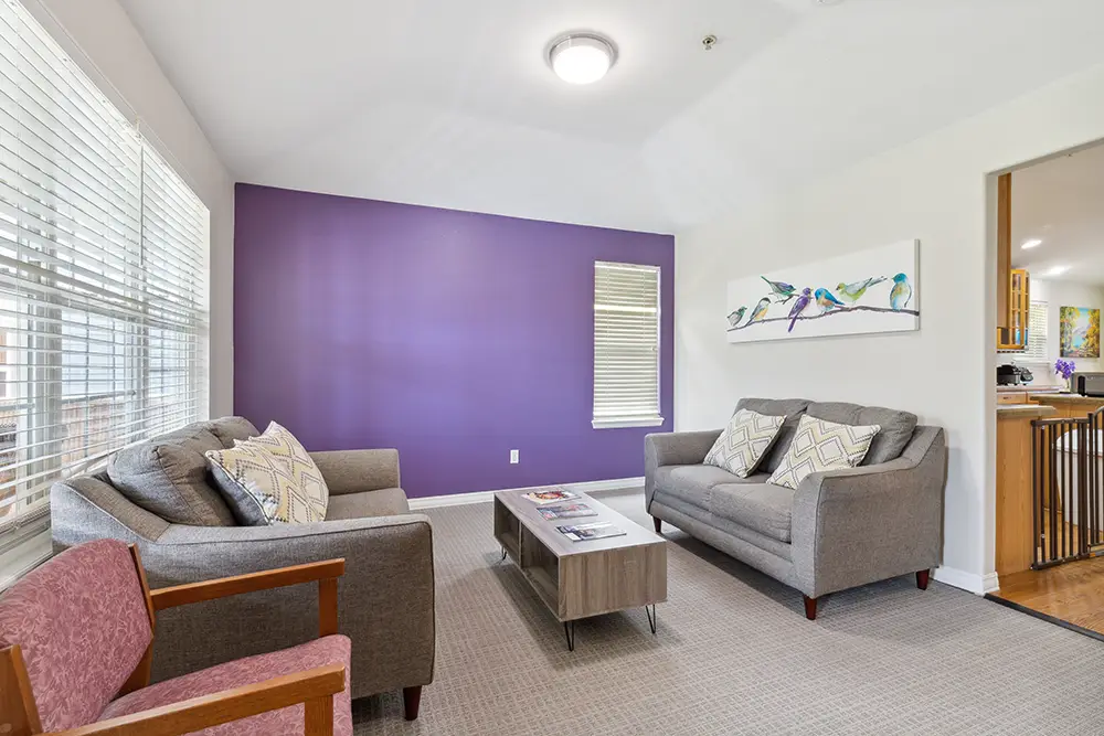Living room with two gray sofas, a wood coffee table, a purple accent wall, a pink armchair, large windows, and a bird artwork above one sofa. The kitchen is visible in the background.