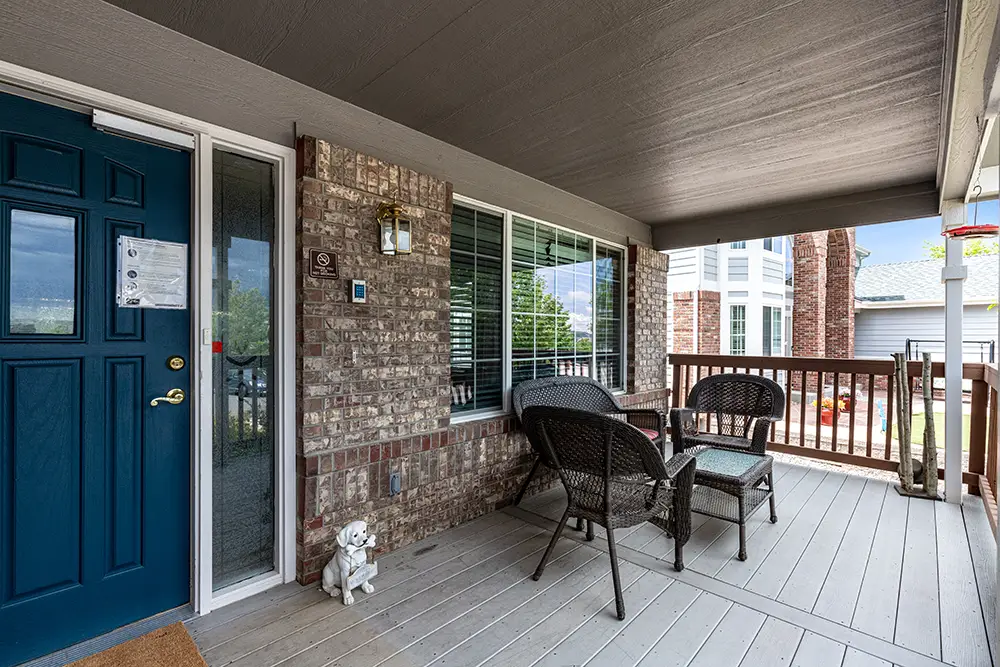 A covered porch with wicker chairs, a small table, a blue front door, brick wall, blinds on the window, and a decorative dog statue near the entrance.