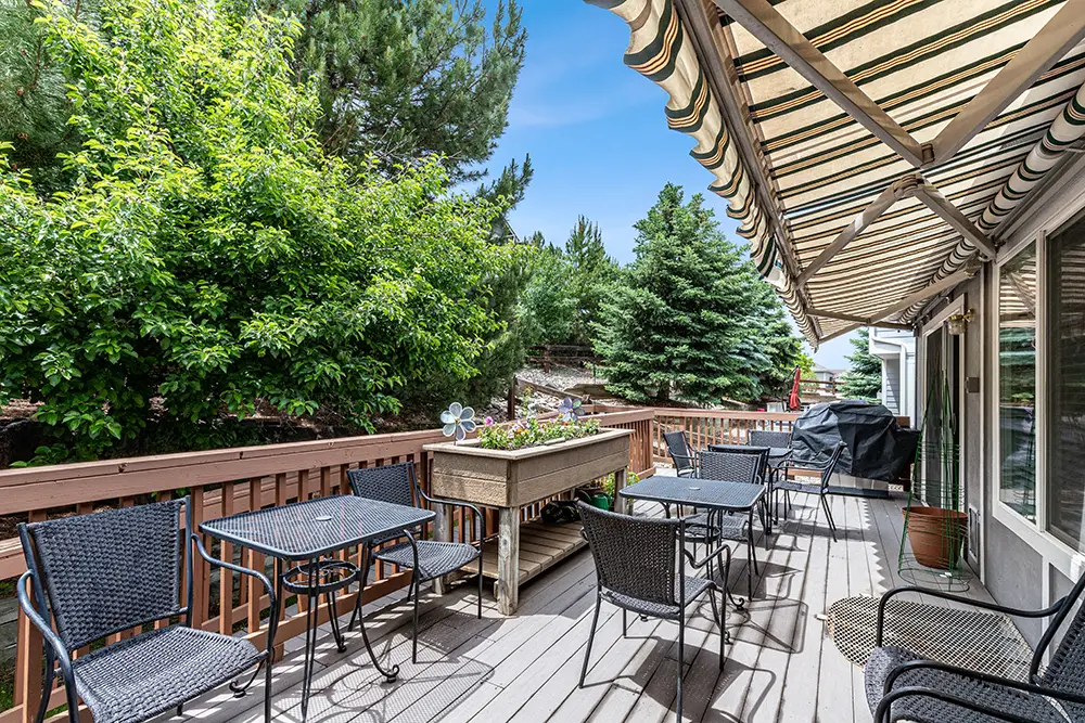 Outdoor deck with black metal tables and chairs, striped awning, planter box, grill, and view of green trees under a blue sky.