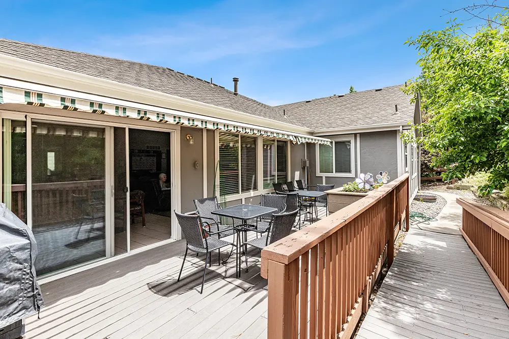 Wooden deck with outdoor dining table and chairs, striped awning, sliding glass doors, and railing next to a pathway and greenery.