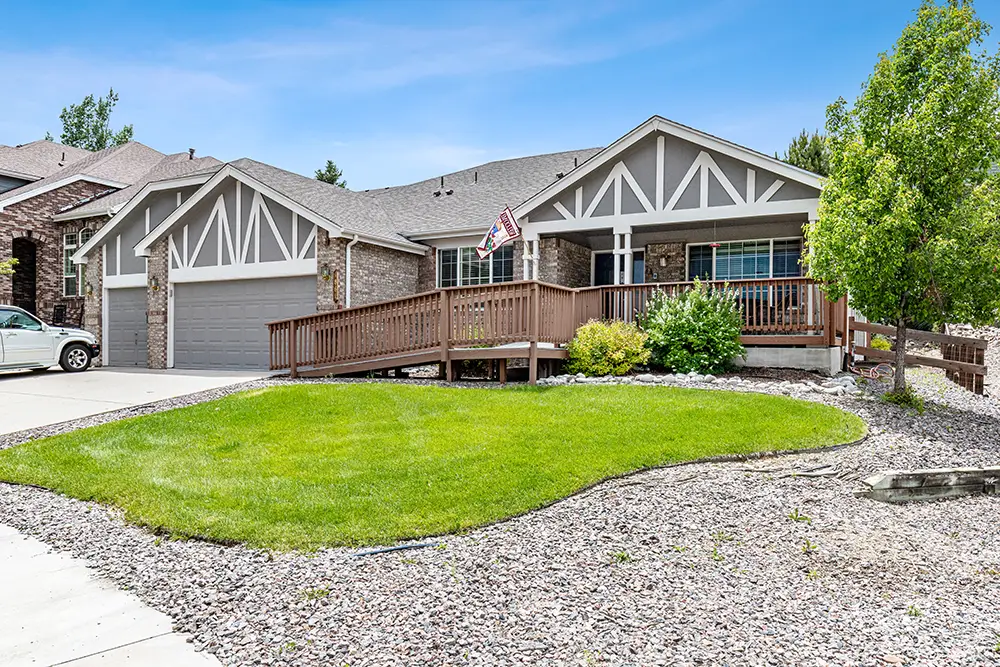 Single-story house with gray siding, a two-car garage, a wooden ramp, green lawn, American flag, and landscaping with rocks under a blue sky.