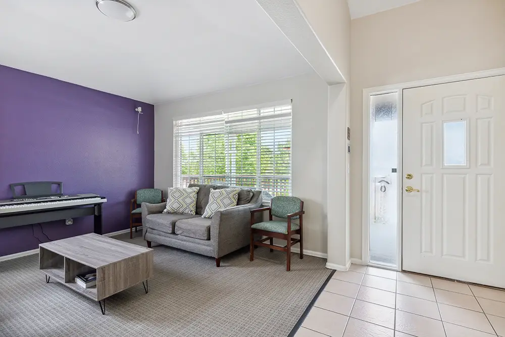 Living room with a purple accent wall, keyboard, gray sofa, two chairs, coffee table, and large window near a white front door with glass side panel.