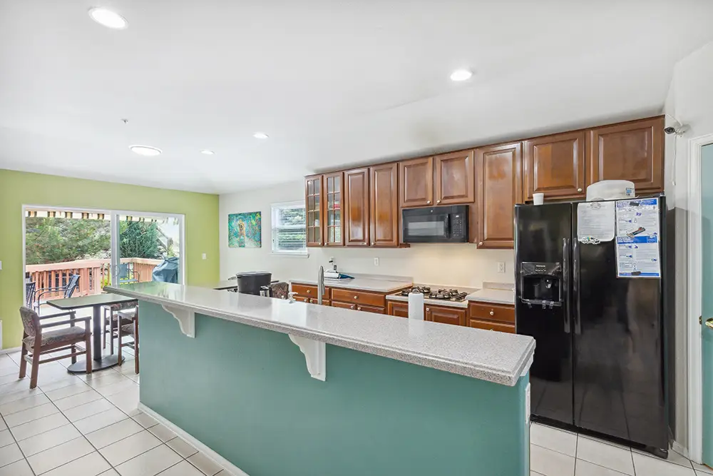 Modern kitchen with brown cabinets, black appliances, and a breakfast bar. Dining area with table and chairs is next to a sliding door opening to a wooden deck with greenery outside.
