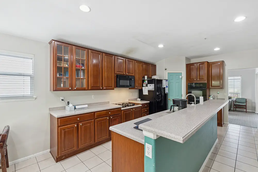 Spacious kitchen with wooden cabinets, black appliances, a long countertop island, white tile floor, and a light blue accent door.