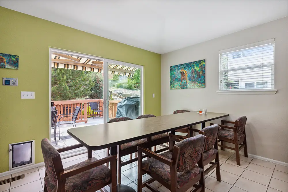 Dining room with a long table, six chairs, tile floor, green accent wall, colorful artwork, and a sliding glass door opening to a deck with outdoor seating and a grill.