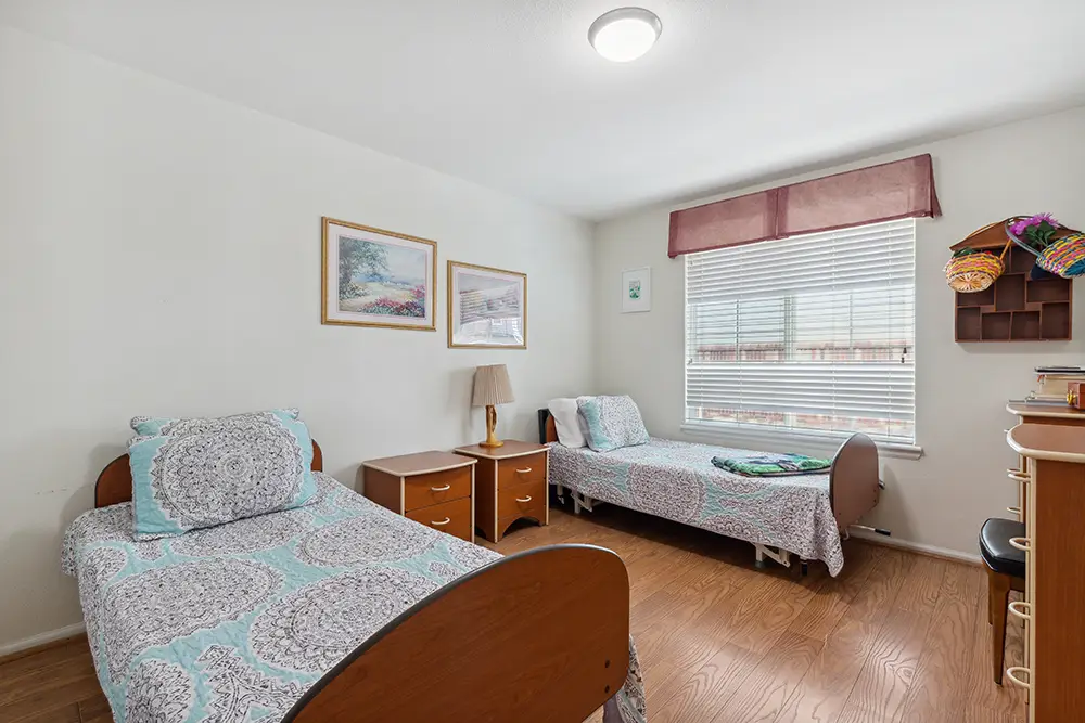 Two single beds with patterned bedding in a tidy, well-lit bedroom featuring wood flooring, a window with blinds, and simple wall art.