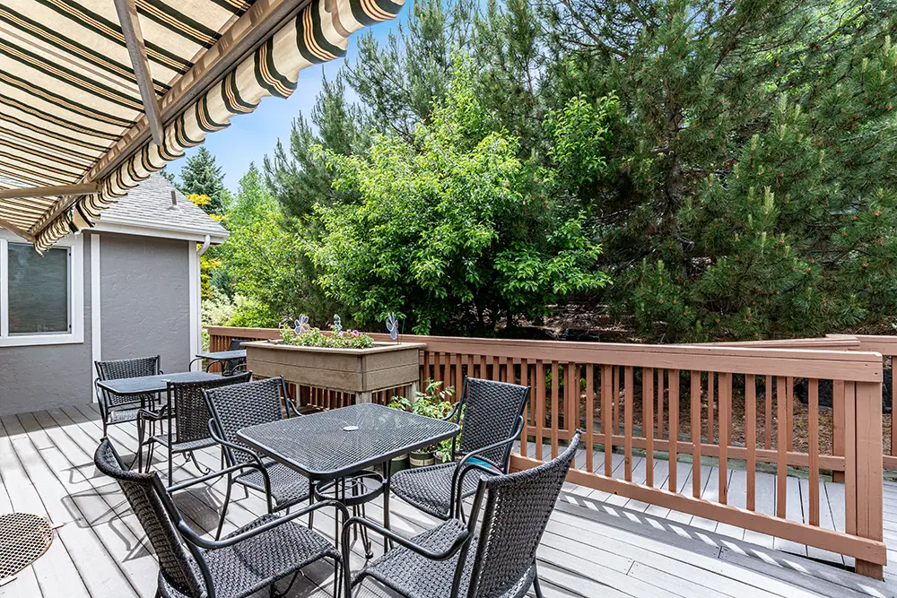 Outdoor patio with black wicker chairs and square tables on a wooden deck, shaded by a striped awning, overlooking trees and greenery.