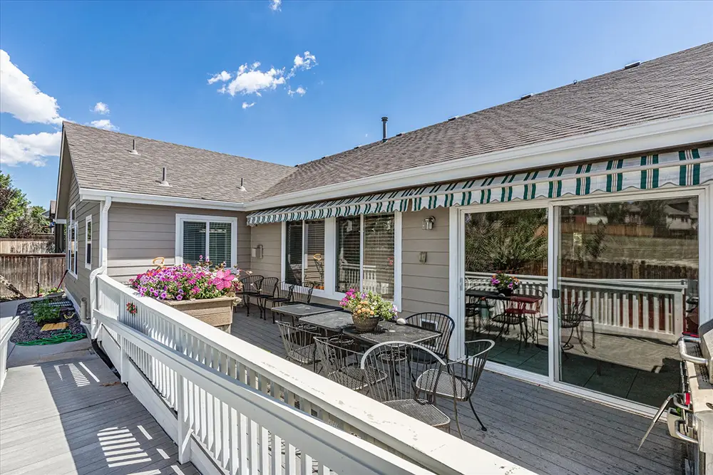 Outdoor wooden deck with tables, chairs, potted flowers, and striped awning attached to a house on a sunny day.