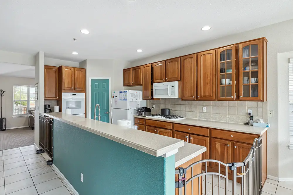 Modern kitchen with wooden cabinets, white appliances, tiled backsplash, and a teal accent wall. A safety gate is installed around the kitchen area, and recessed lighting is on the ceiling.