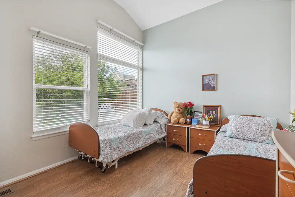 Two twin beds with patterned bedding in a bright room with large windows, a shared nightstand, a teddy bear, flowers, framed photos, and wood flooring.