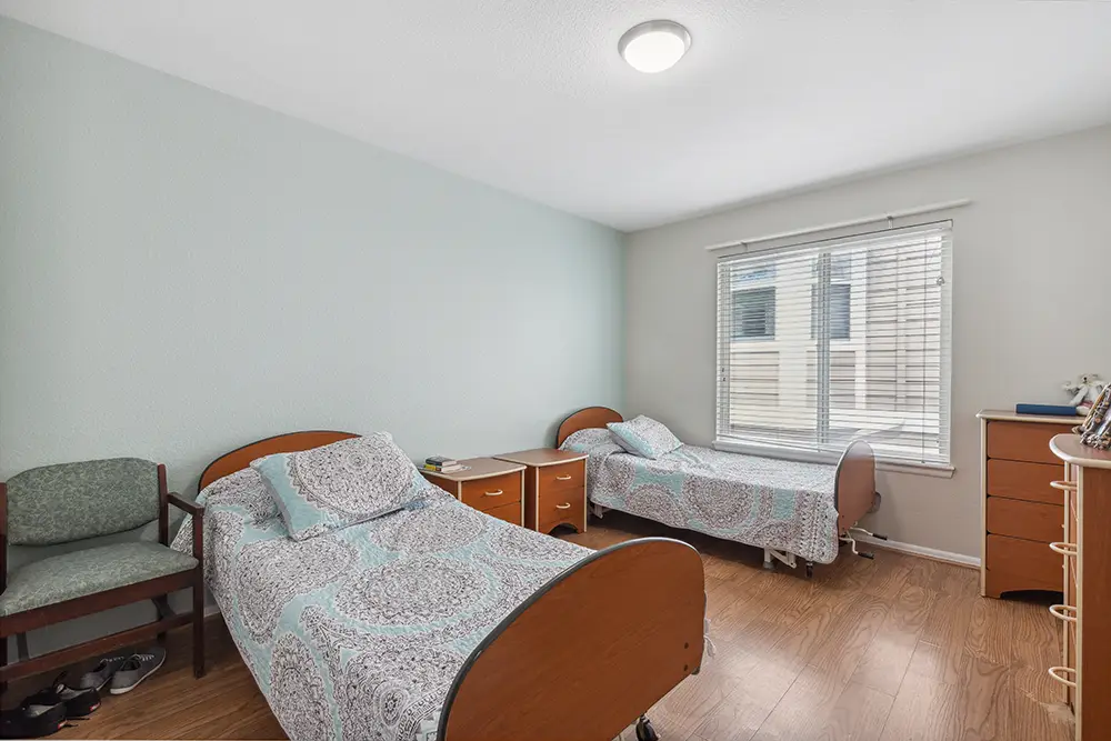 A simple bedroom with two twin beds, a nightstand, a dresser, a green chair, and a window with blinds letting in natural light.