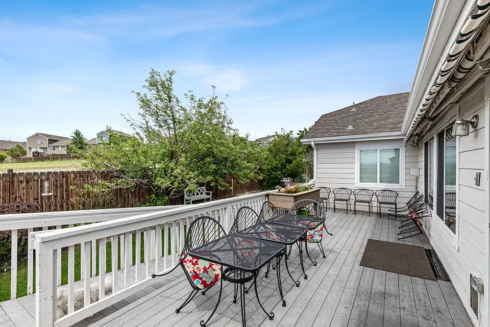 A spacious backyard deck with a black metal table, six chairs with floral cushions, a wooden railing, and a view of a grassy yard and neighboring homes under a clear blue sky.