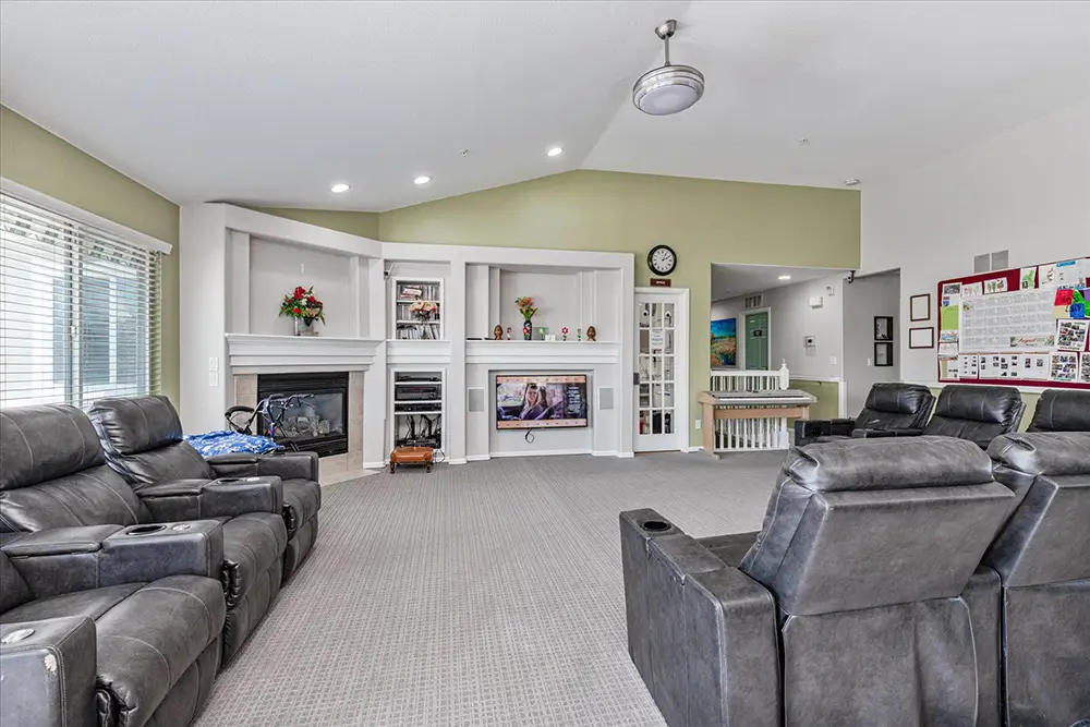 Spacious living room with gray recliners, wall-mounted TV, fireplace, large windows, and a bulletin board on the right wall.