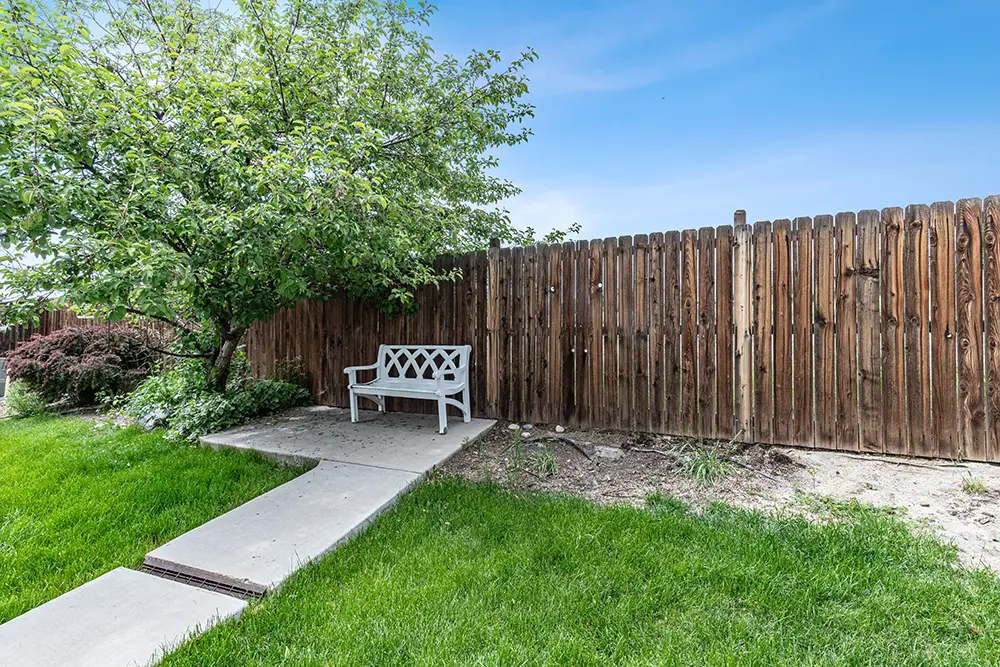 A white bench sits on a concrete path near a wooden fence and a leafy tree, surrounded by green grass and some bare soil patches.