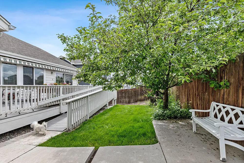 Accessible backyard with a white ramp, green grass, a leafy tree, wooden fence, white bench, and a small dog walking near the ramp.