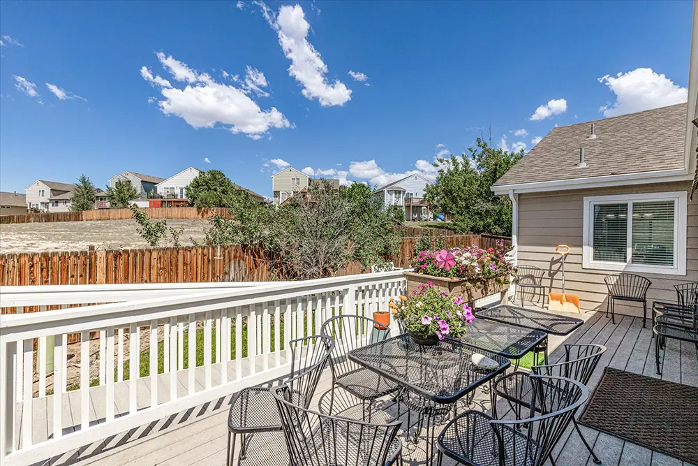 Outdoor patio with metal tables and chairs, potted flowers, and a wooden fence in a suburban backyard under a blue sky with scattered clouds.
