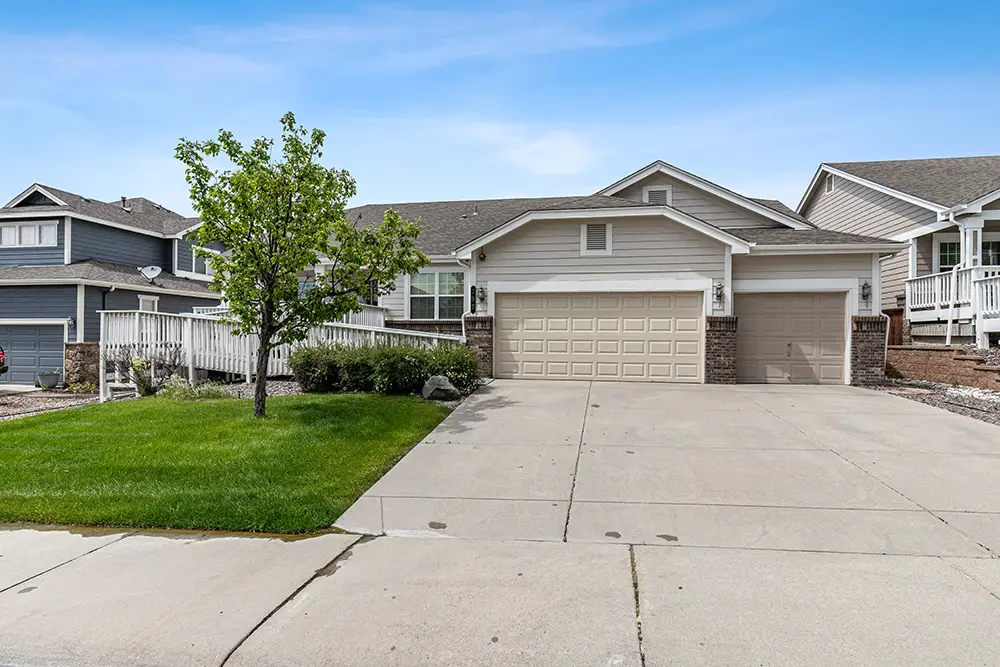 Single-story suburban house with a three-car garage, small front lawn, and a young tree, set under a clear sky.