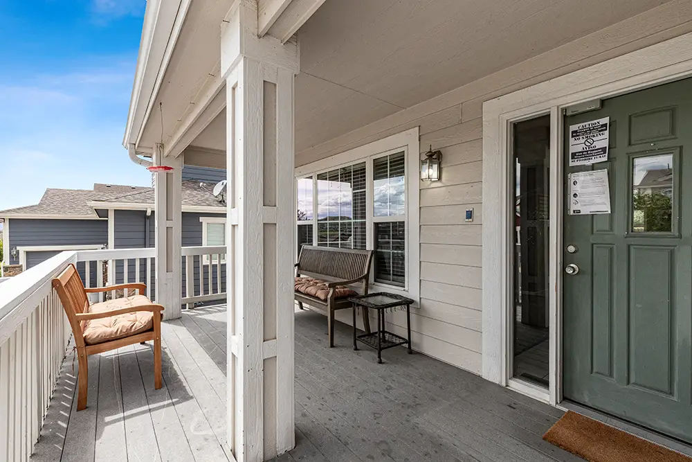 Covered front porch with a green door, bench, and table; wood railing and siding; window with blinds; another bench and homes visible in the background.