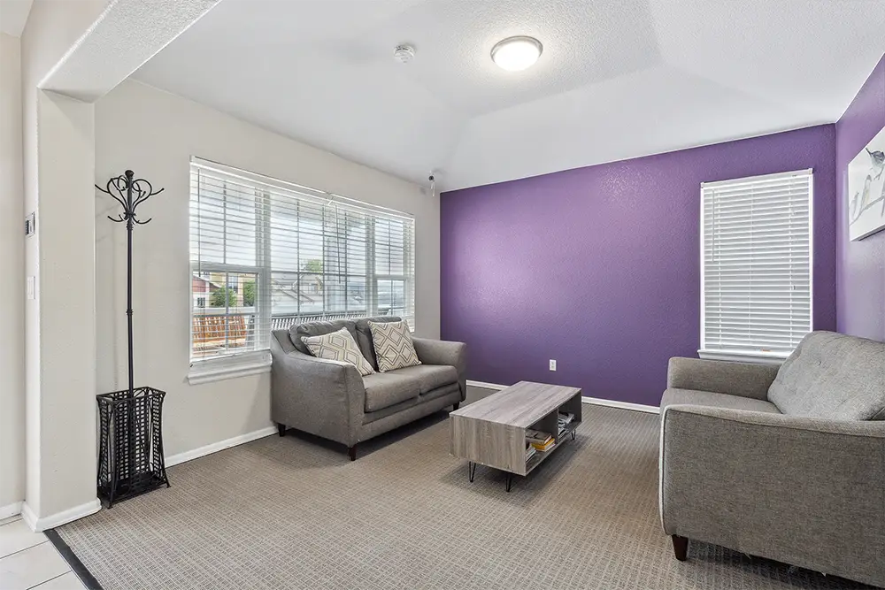Living room with two gray sofas, a wooden coffee table, and a purple accent wall, next to windows with blinds and a standing coat rack.