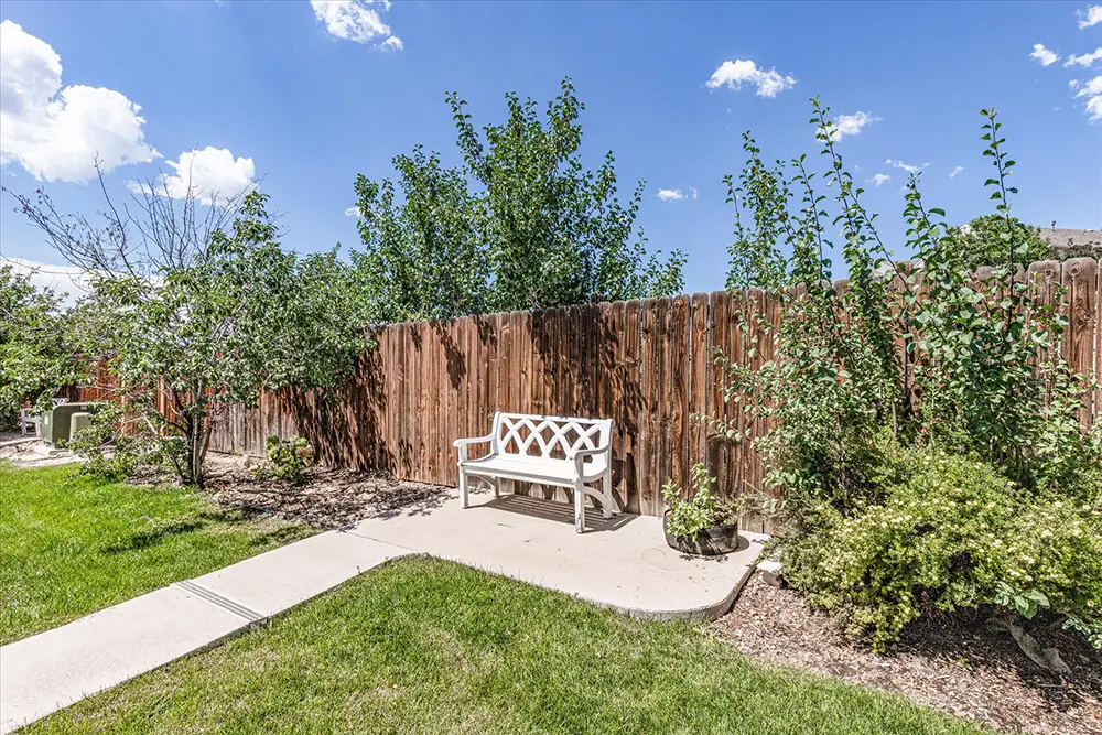 A white bench sits on a concrete path next to a wooden fence, surrounded by green grass, bushes, and trees under a blue sky with clouds.