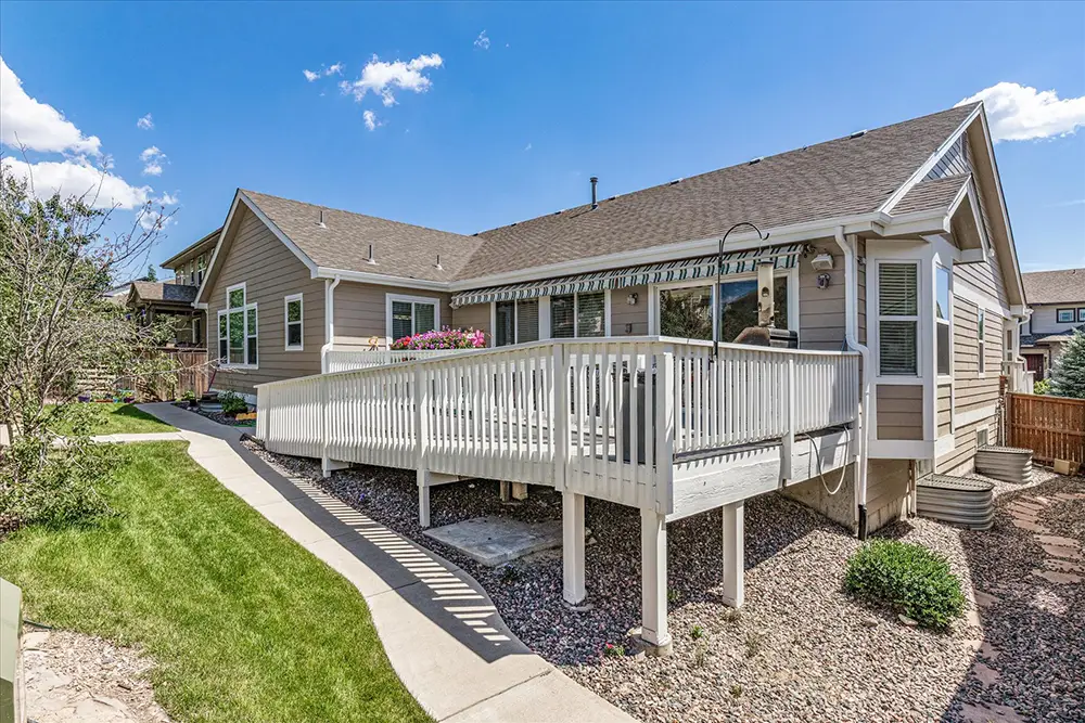 Single-story house with beige siding, a gable roof, and a white ramp leading to a raised deck. The yard features grass, shrubs, a stone border, and a clear, sunny sky.