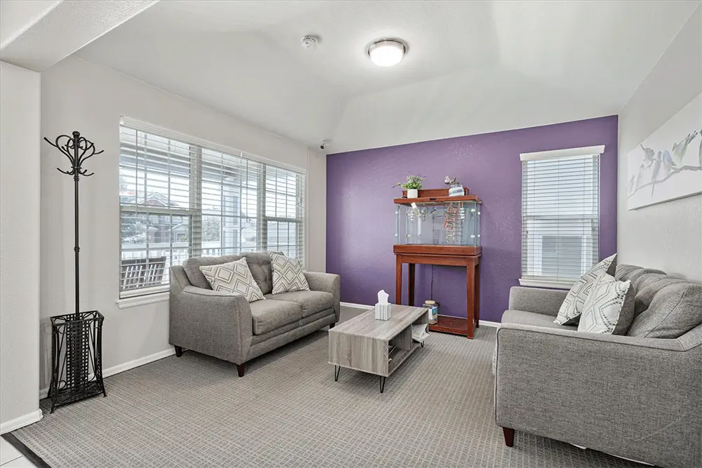Living room with two gray sofas, a wooden coffee table, a fish tank on a stand, and a purple accent wall, featuring large windows and neutral decor.