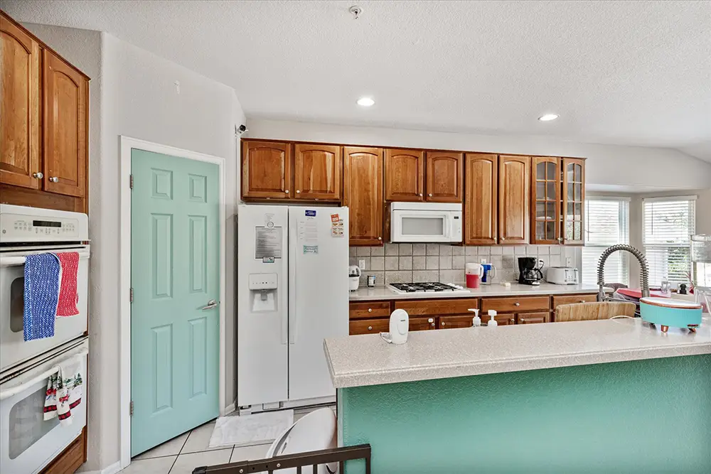 Bright kitchen with wooden cabinets, white appliances, a mint green door, and a light green breakfast bar. Countertop holds a dish rack, faucet, and small appliances.