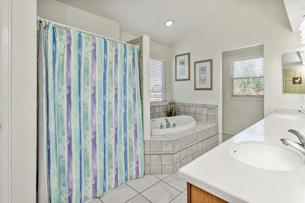 Bathroom with dual sinks, a striped shower curtain, a corner bathtub with tile surround, white countertops, framed art, and two windows with blinds.