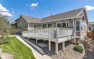 Single-story house with beige siding in Castle Rock, featuring a backyard deck with white railing and awning, sloped lawn, and stone landscaping—an inviting setting ideal for Memory Care needs on a sunny day.