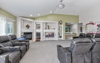 Spacious living room in Castle Rock with gray recliners, a fireplace, wall-mounted TV, large windows, and a bulletin board on the right wall—ideal for Memory Care or TBI Support residents to relax and connect.