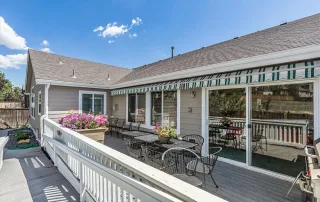 A wooden deck in Castle Rock with patio tables and chairs, flower planters, and a striped retractable awning offers a welcoming space—perfect for Memory Care or TBI Support residents to relax outdoors at this light-colored house.