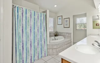 Bathroom with a striped shower curtain, tiled bathtub, double sinks, white countertops, and two windows with blinds—ideal for Memory Care needs in Castle Rock.