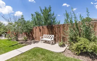 A white wooden bench sits on a concrete path next to a wooden fence in Castle Rock, surrounded by green grass, shrubs, and trees under a partly cloudy sky.