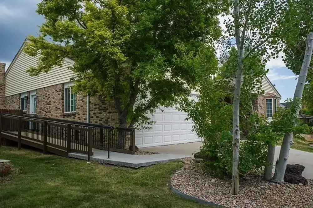 Single-story brick house with attached garage, ramp leading to front entrance, and trees and rocks in landscaped front yard.