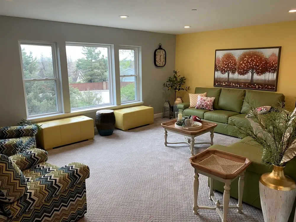 Living room with green sofa, patterned armchairs, large windows, yellow accent wall, tree artwork, and various decorative items on tables and walls.