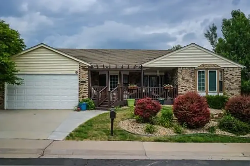 Single-story house with brick and siding exterior, attached two-car garage, front porch, landscaped yard, and driveway under a cloudy sky.