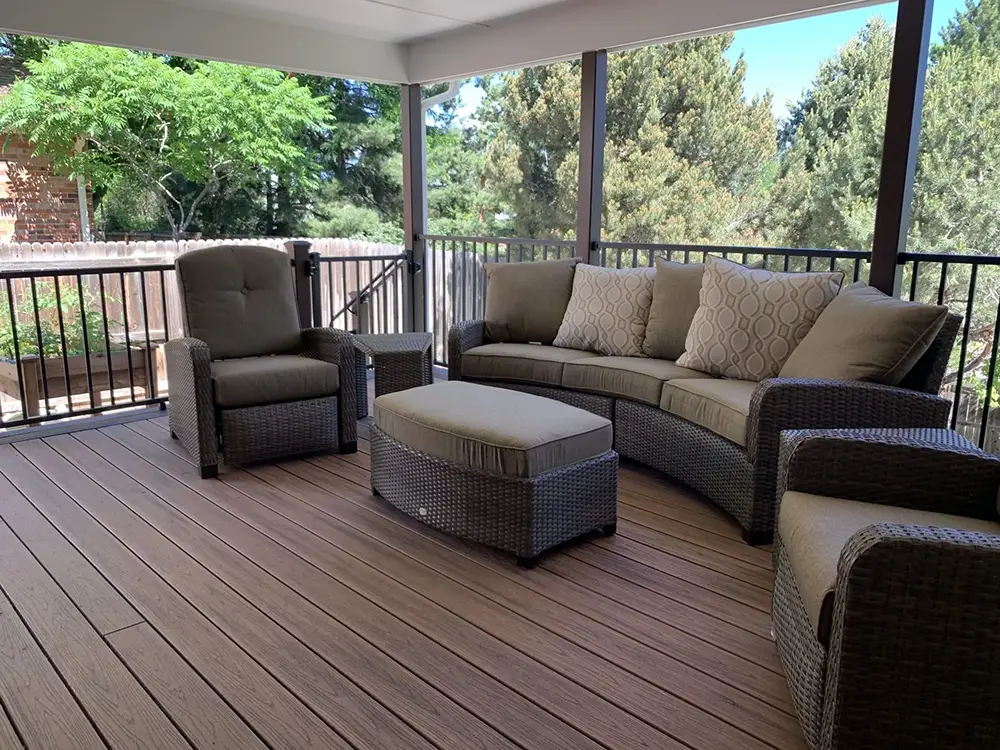 Outdoor patio with brown wicker furniture, including a sofa, two armchairs, a cushioned ottoman, and a view of trees through a railing.
