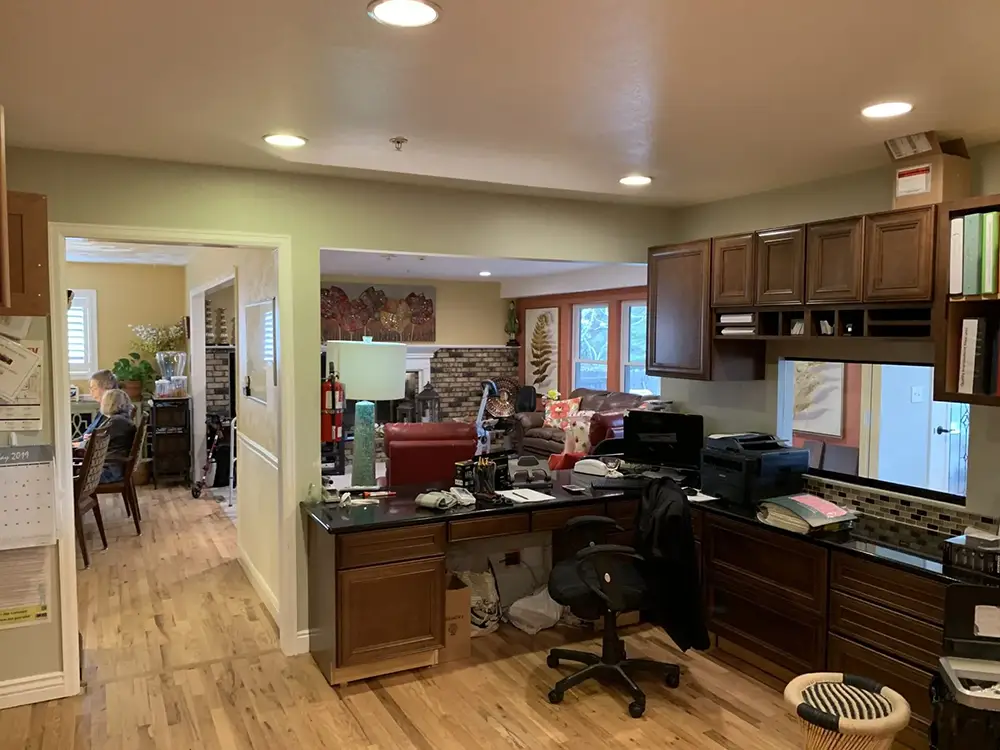 Office area with multiple desks, chairs, and computers; a person is seen working in an adjacent room. The space has wooden floors, brown cabinets, and overhead lights.