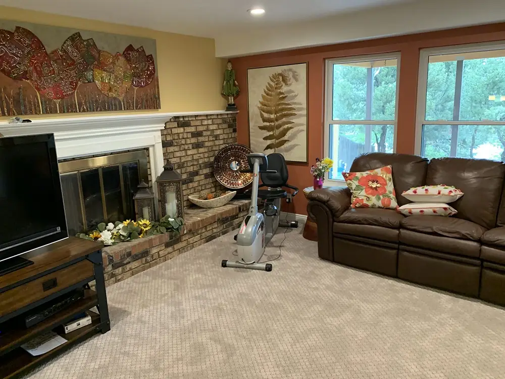 Living room with brown leather couch, TV, exercise bike, fireplace with brick surround, and large windows; decorated with wall art and cushions.