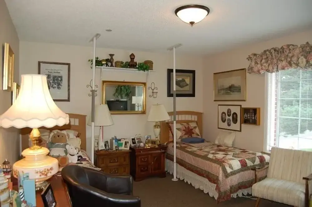 A tidy bedroom with two twin beds, a desk, lamp, chairs, framed art on the walls, and sunlight coming through a window with floral curtains.