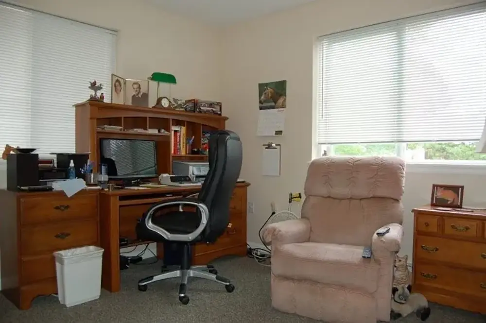 Home office with a desk, computer, black chair, beige recliner, two windows with blinds, framed photos, a lamp, and a small wooden cabinet.