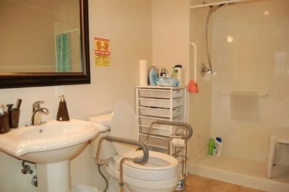 Accessible bathroom with a raised toilet seat and grab bars, a pedestal sink, a walk-in shower with a fold-down seat, and a storage unit with toiletries.