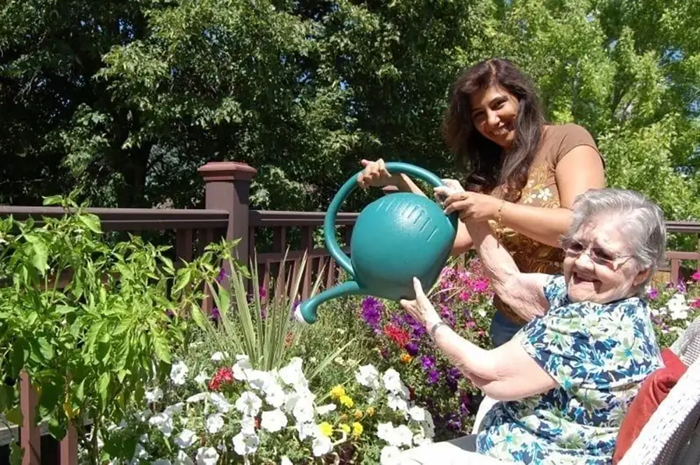 An older woman and a younger woman water flowers together on a patio surrounded by green plants and trees on a sunny day.