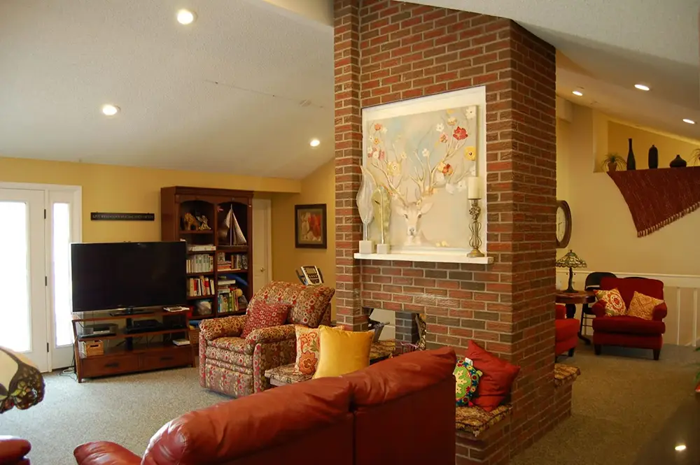 Living room with red sofas, floral armchairs, a brick fireplace, TV, shelves, and wall art above the mantel; warm lighting and decorative pillows.