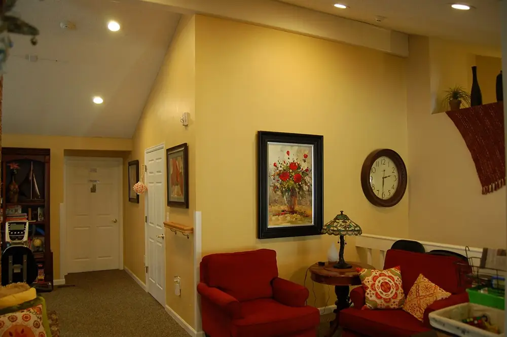 A living room with yellow walls, two red chairs, a floral painting, a wall clock, a decorative lamp, and patterned pillows.
